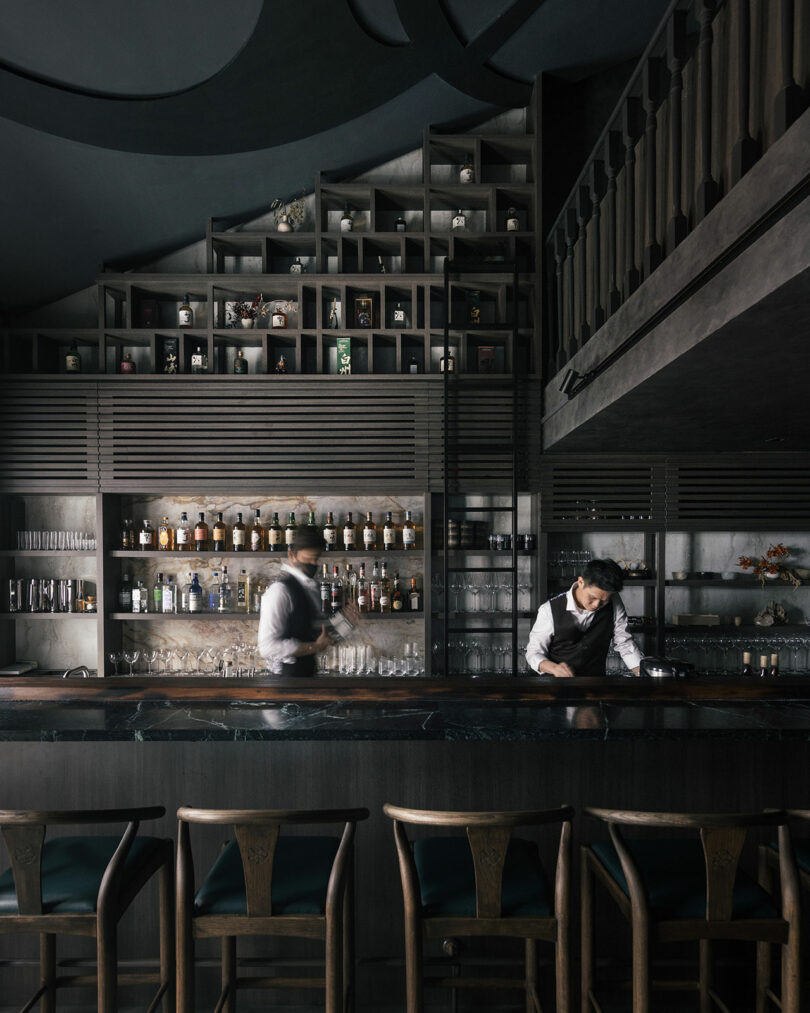 Dimly lit, Amanda Gunawan-designed modern bar with two bartenders expertly preparing drinks. Elegant shelves of bottles and glassware adorn the background, while sleek wooden stools invite guests to unwind in front of the stylish bar.