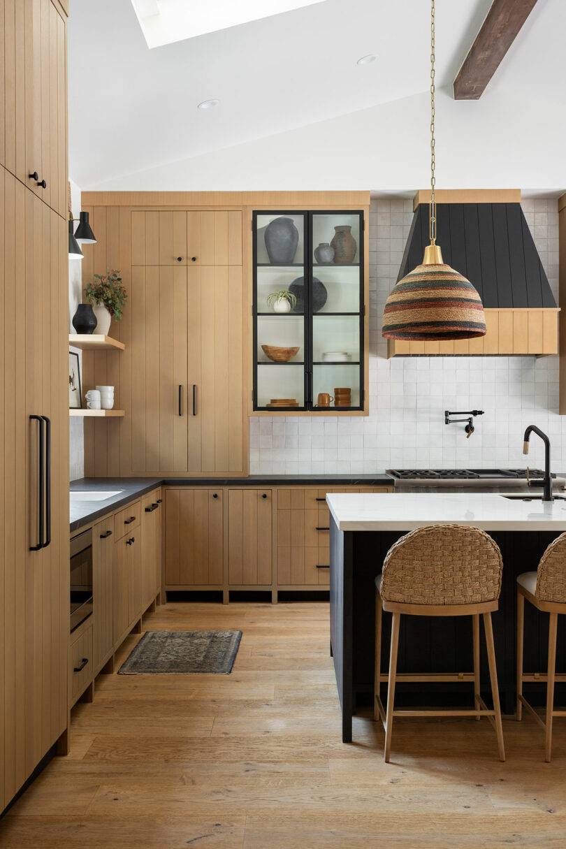 Modern kitchen with wooden cabinets, island with woven chairs, pendant light, and dark accents. Cabinets display various dishes. Natural light from skylight brightens space.