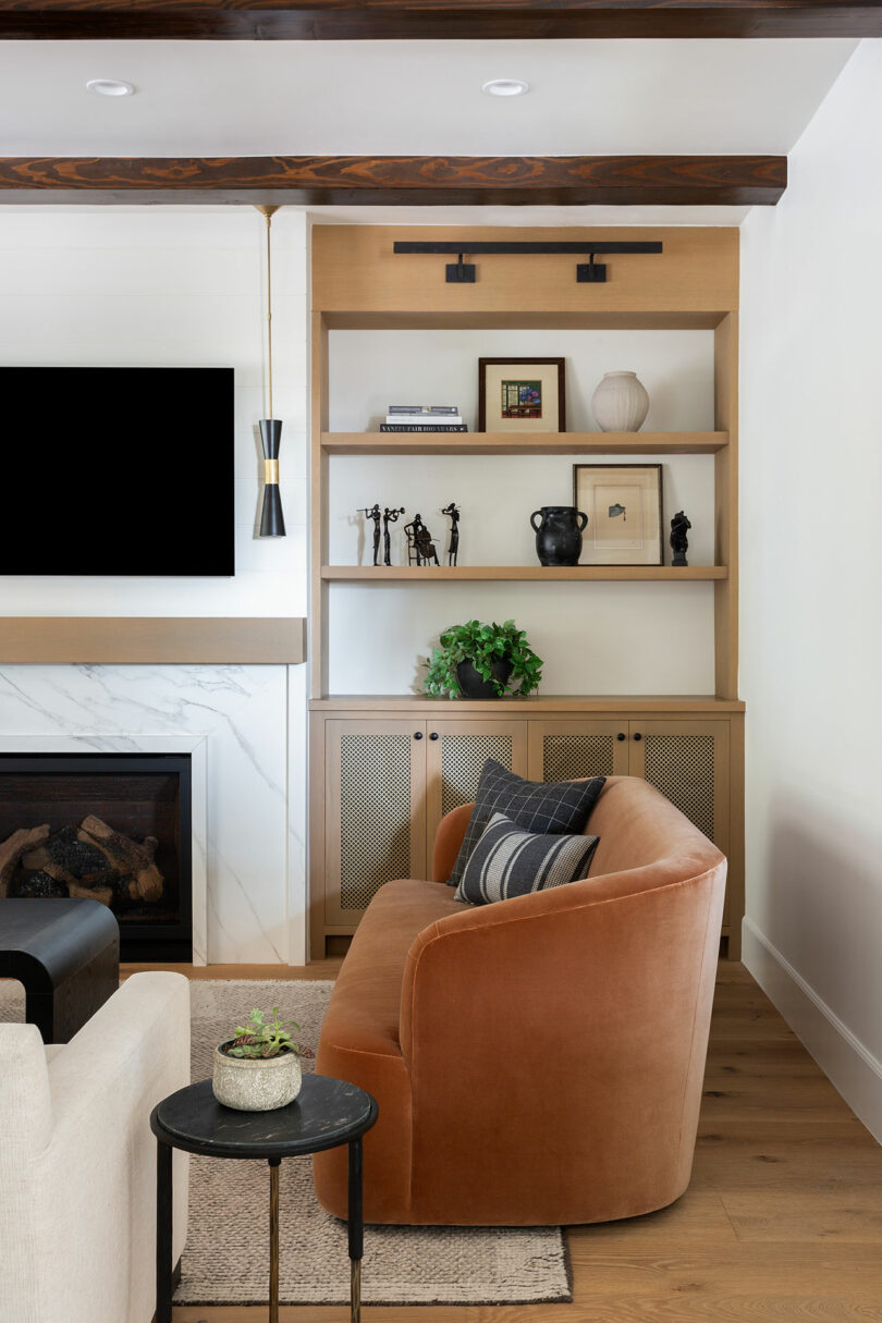 Living room with a brown curved sofa, black side table, and built-in shelves. Shelves hold decorative items and plants. A modern fireplace and wall-mounted TV are on the adjacent wall.