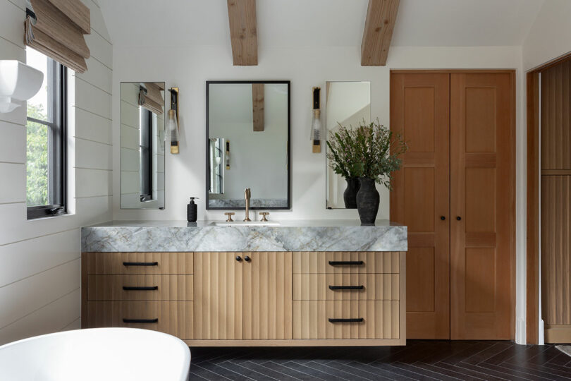 Modern bathroom with double vanity, marble countertop, two mirrors, wooden cabinets, and wall lights. Black herringbone floor and vase with greenery on the counter.