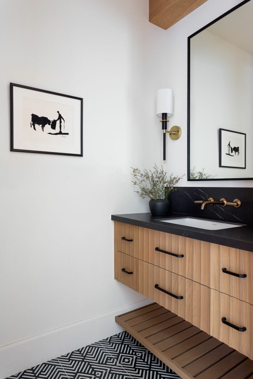 A modern bathroom features a black countertop, a wooden vanity with a large mirror, a wall sconce, a potted plant, and a framed silhouette artwork of a person with a bull.