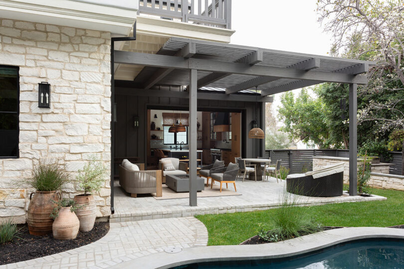 Outdoor patio with a dark wood pergola, beige seating, a dining table, potted plants, and a pool in the foreground. The patio opens into a house with large windows and stone walls.