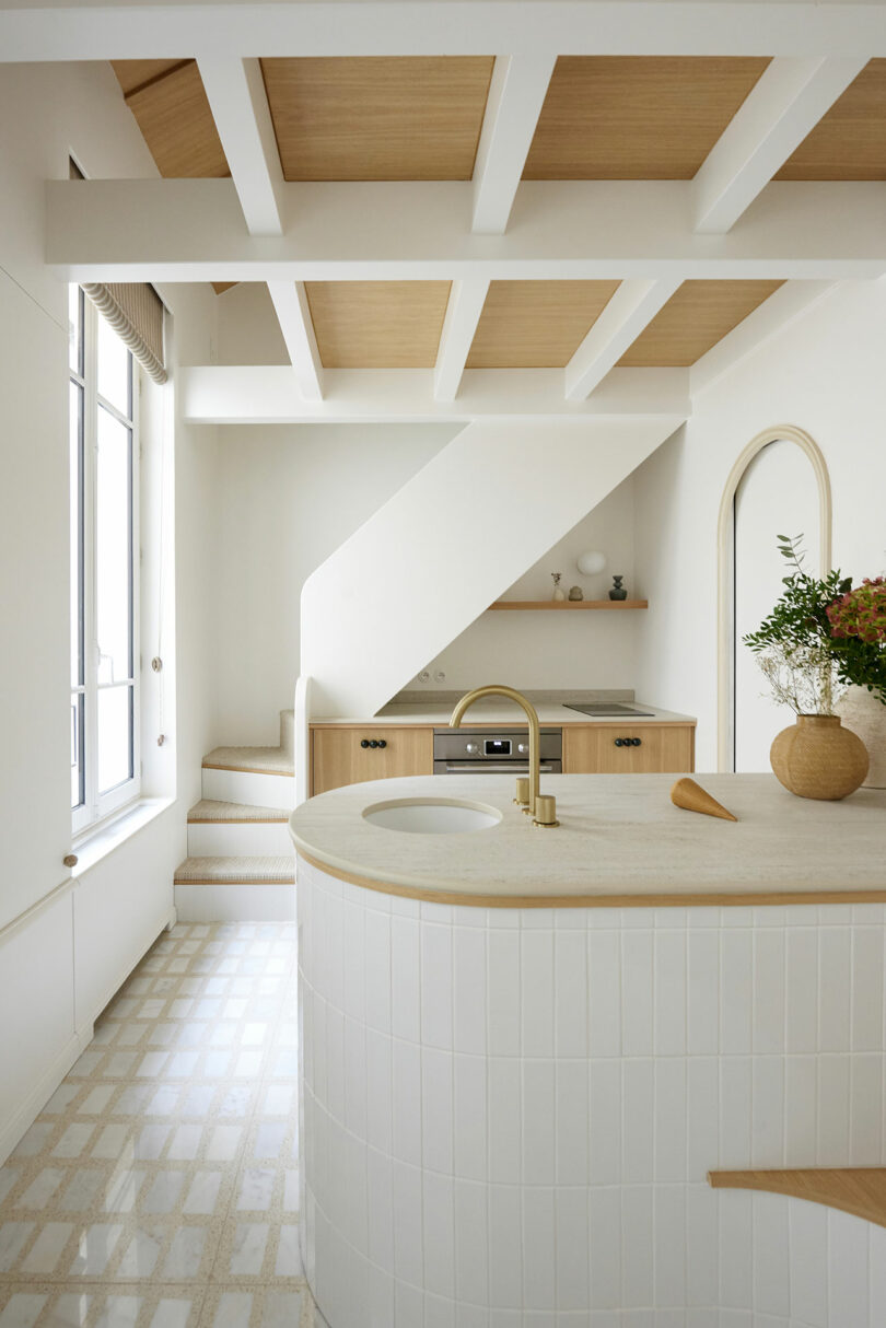 Modern kitchen with white tile and wood accents, featuring a curved island with a sink, plants on the counter, and an oven under the stairs. Stairs lead up, and a large window lets in natural light.