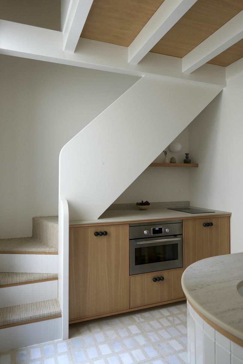 Modern kitchen with wood cabinetry under a staircase, featuring a built-in oven, minimalist decor on a shelf, and tiled flooring.