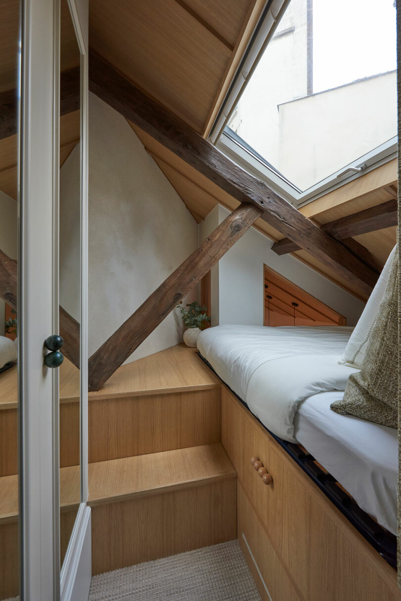 Cozy attic bedroom with wooden beams and a skylight. Features a built-in bed with white bedding and steps leading up, surrounded by light wood paneling and a small plant decoration.