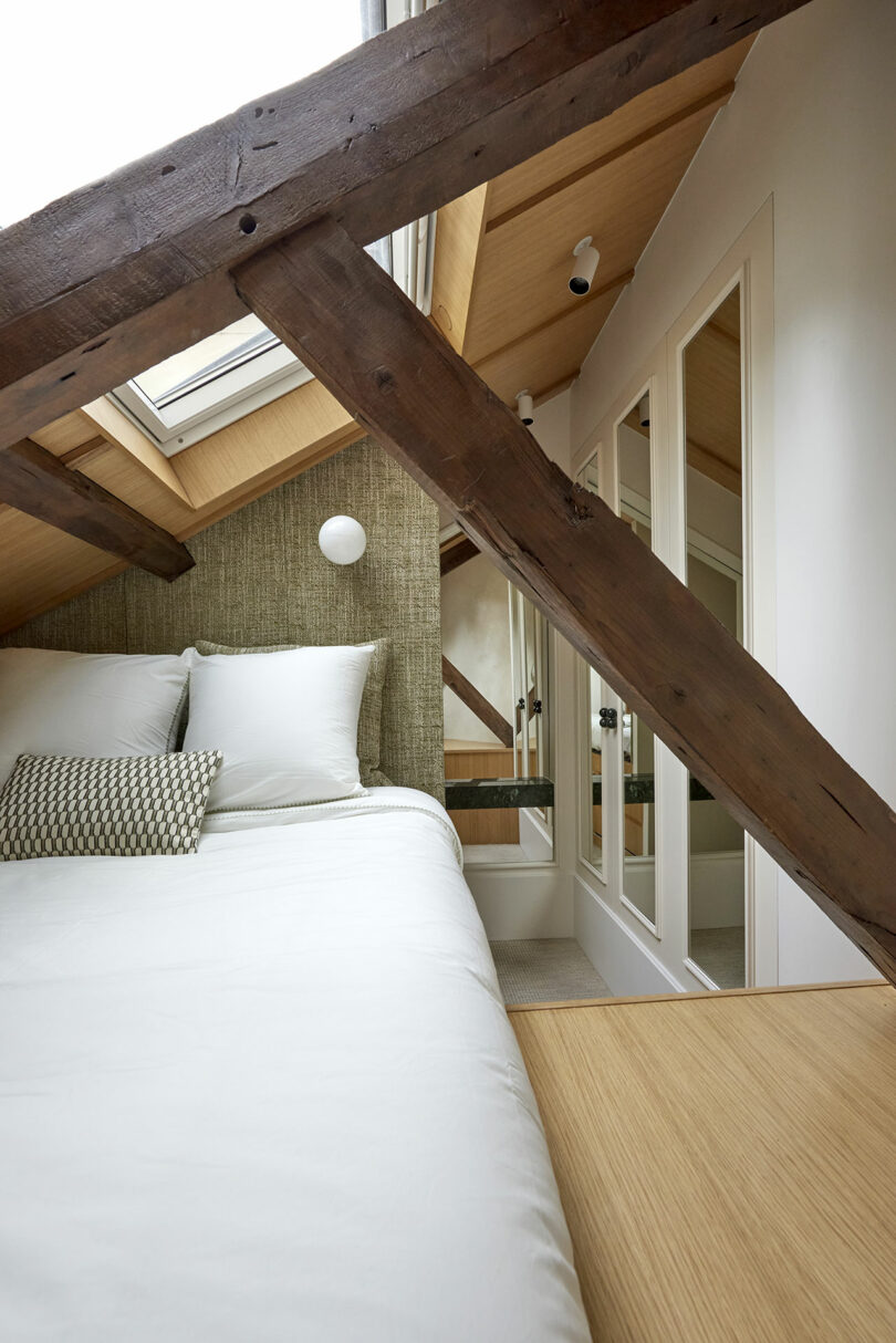 Attic bedroom with exposed wooden beams, a skylight, white bedding on the bed, and a mirrored wardrobe on the right.