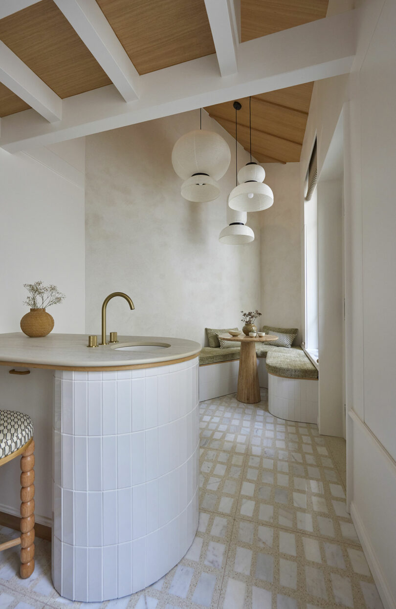 Modern kitchen with white tiled island, brass faucet, and wood accents. Overhead, three pendant lights hang. A cozy nook with green cushioned seating and small wooden table is in the background.