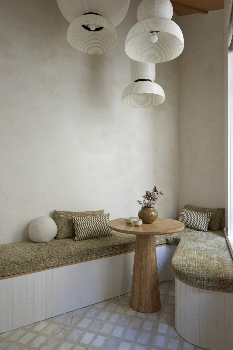 Cozy seating nook with cushioned benches, a small round wooden table, a vase with flowers, and hanging paper lanterns. Neutral colors and soft lighting create a serene atmosphere.