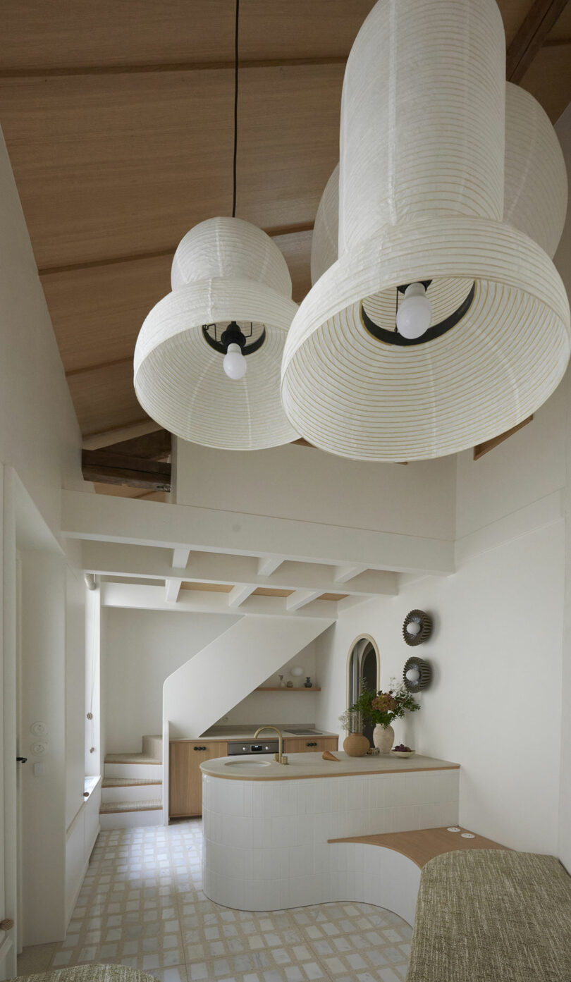 Minimalist kitchen with white curved island, wooden ceiling, and large white pendant lights. A staircase leads upstairs, and a small plant sits on a countertop.