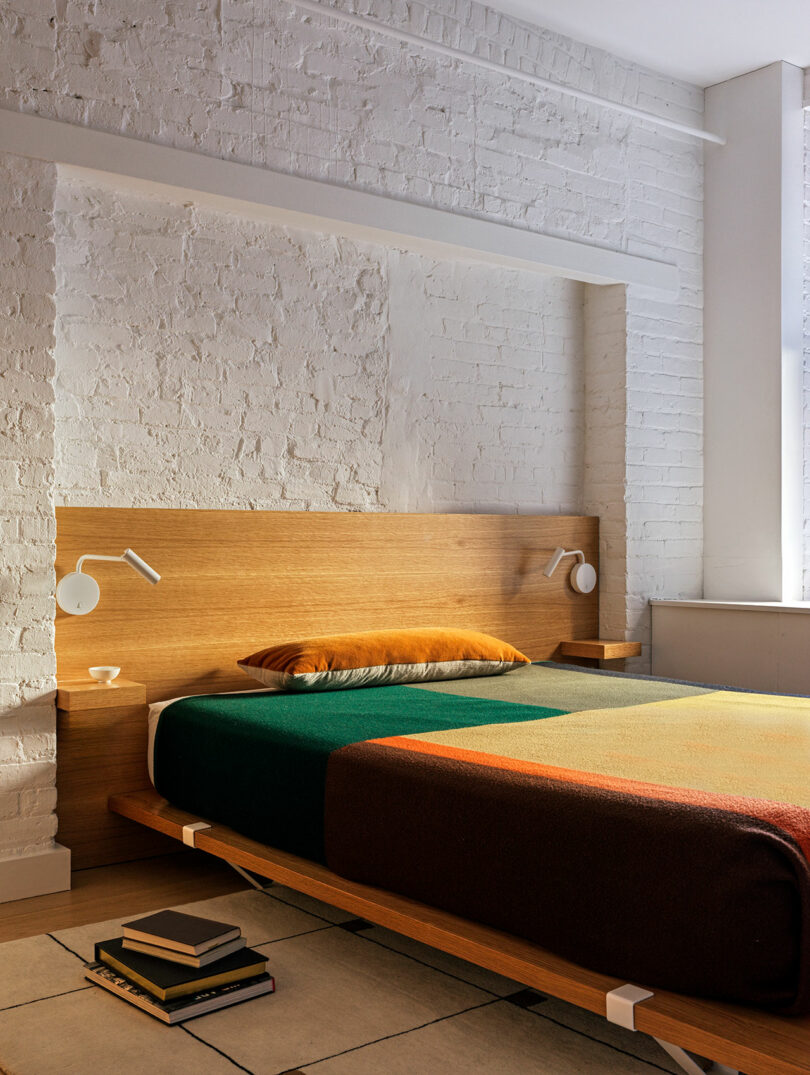 A modern bedroom features a wooden bed with a multicolored blanket, two wall-mounted lamps, a brick wall backdrop, and books stacked on the floor.