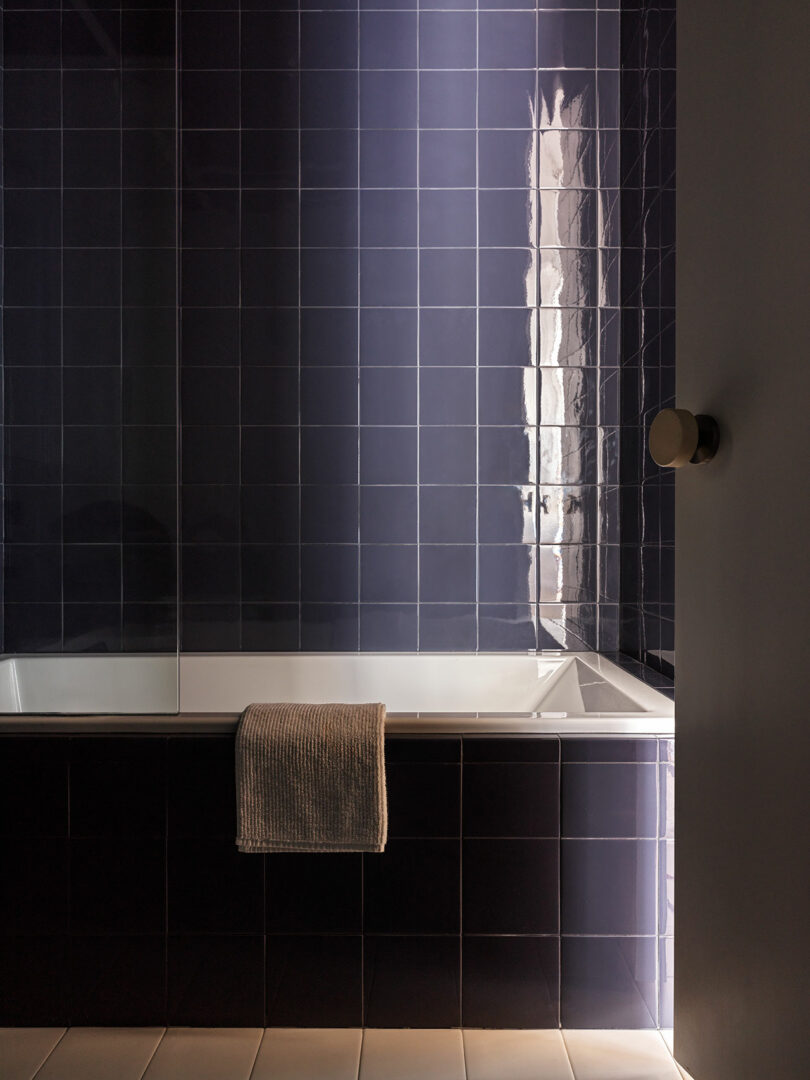 A bathroom with dark blue tiled walls and a bath. A beige towel hangs over the edge of the tub. Light casts a reflection on the tiles.