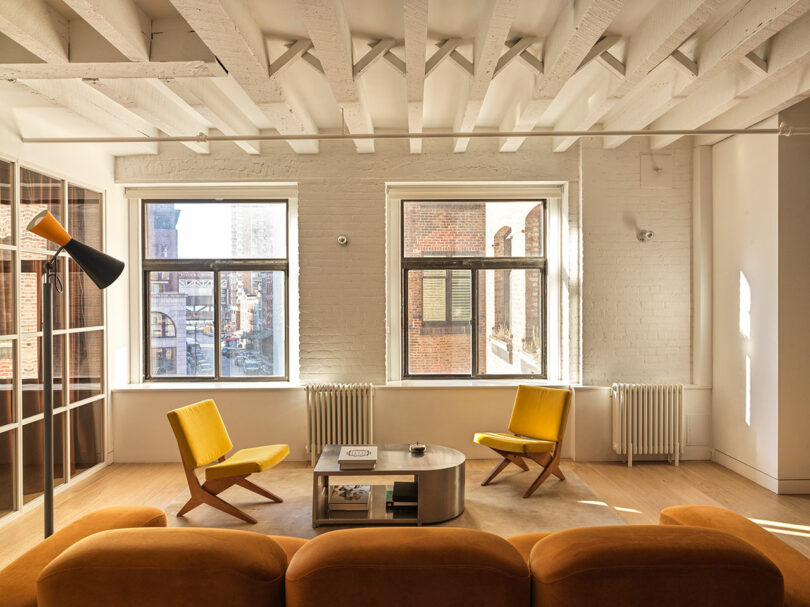A modern living room with two yellow chairs, a brown sofa, a gray coffee table, and a large black and orange floor lamp. Sunlight enters through three windows, illuminating the wood floor.