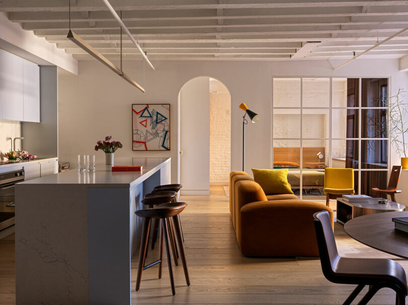 A modern living space with an open kitchen, wooden floors, brown sofa, colorful cushions, and artwork on walls. A large window and ceiling beams enhance the room's brightness.