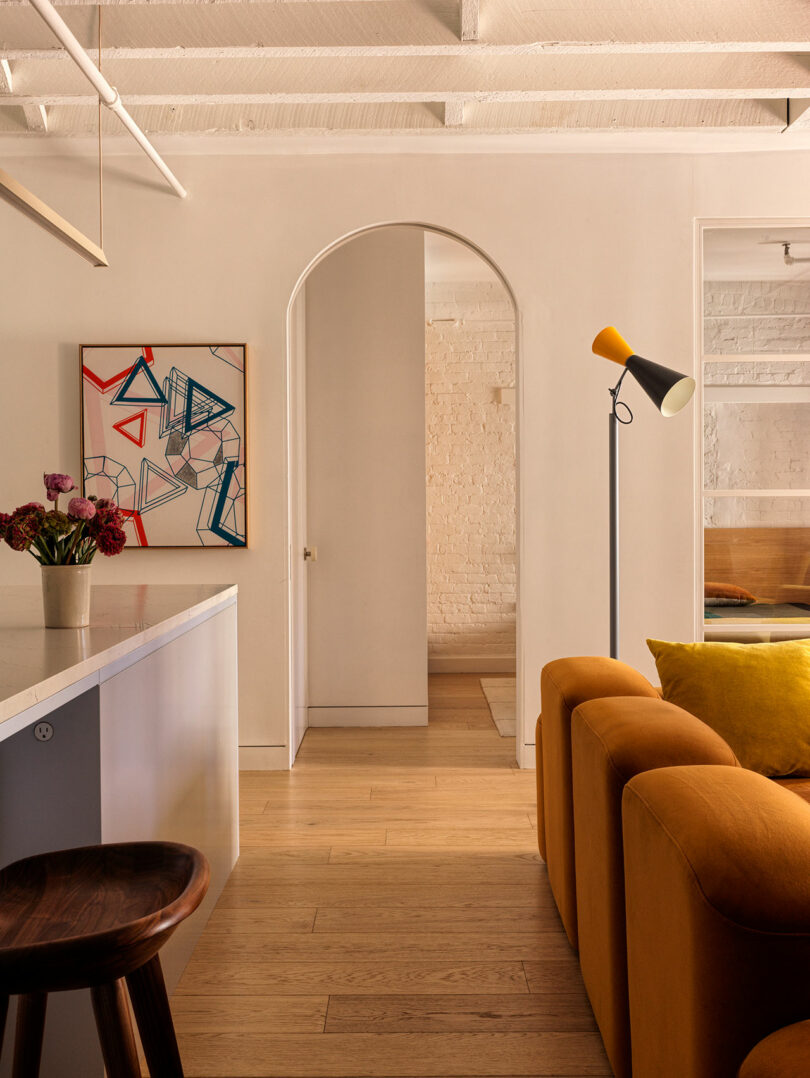 Living room with an archway, mustard sofa, modern artwork on the wall, and a standing lamp. Hardwood flooring and exposed ceiling beams enhance the space's warmth and texture.