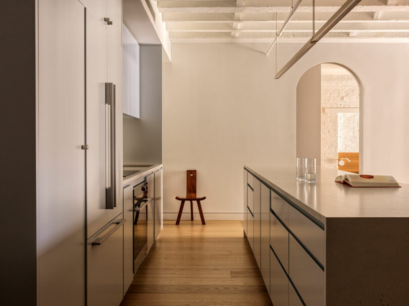 Minimalist kitchen with sleek cabinetry, island countertop, and wooden flooring. A narrow chair sits by an arched doorway. Open book and two glasses on the island. White ceiling beams above.