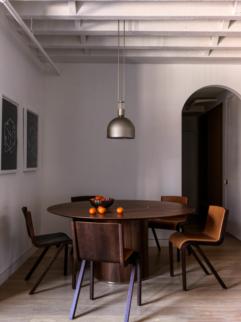 A modern dining room with a round wooden table and four chairs. A pendant light hangs above the table, which holds a bowl and oranges. Paintings adorn the white wall, and an archway leads to another room.