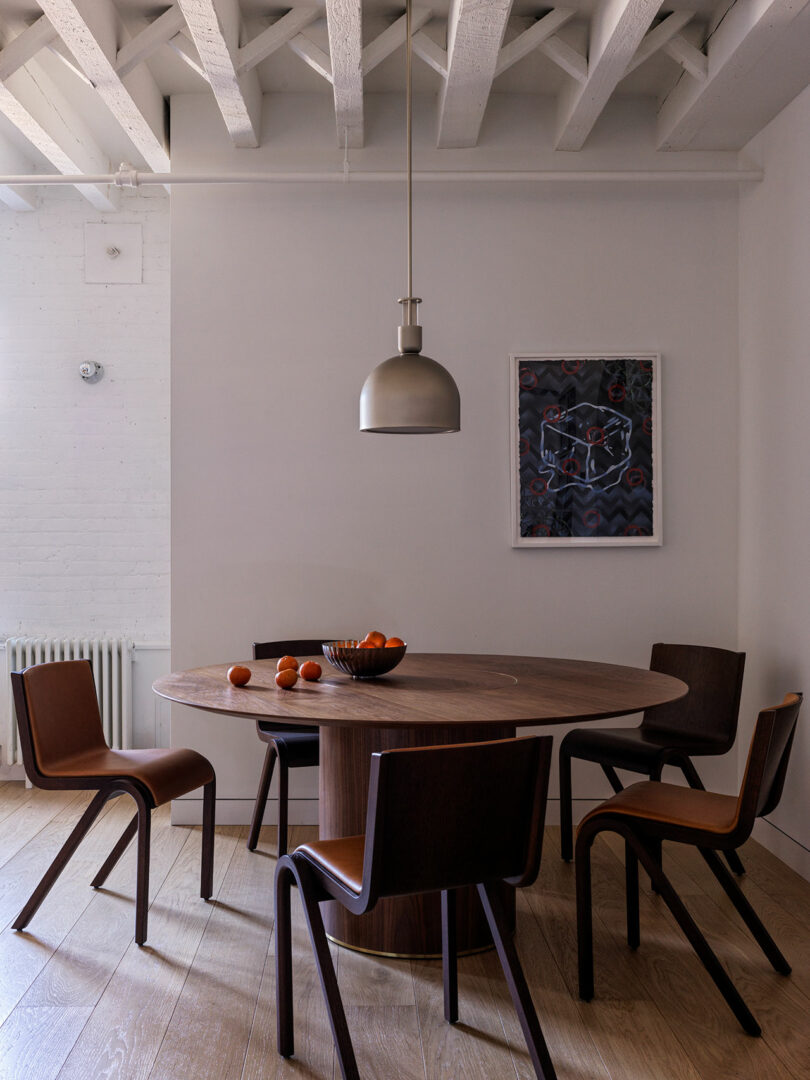 Modern dining room with a round wooden table, four brown chairs, and a pendant light. A bowl of oranges and scattered fruits on the table. Art on the wall and exposed ceiling beams above.
