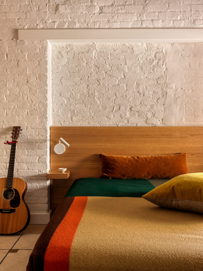 A neatly arranged bed with colorful pillows and a blanket, next to a small wooden shelf holding a lamp and mug, with a guitar leaning against a white brick wall.