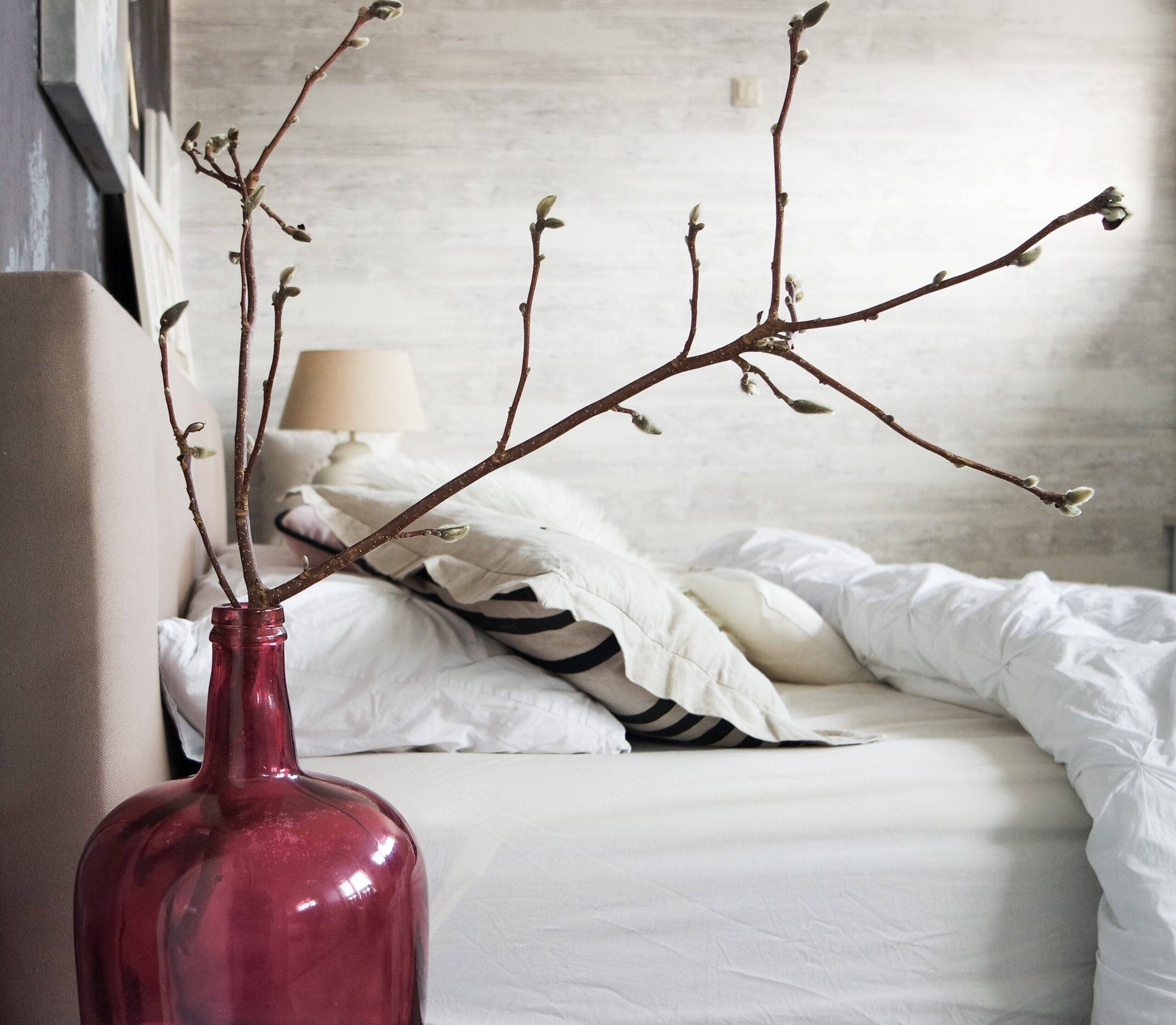 A red vase with some large decorative branches in it next to a bed.