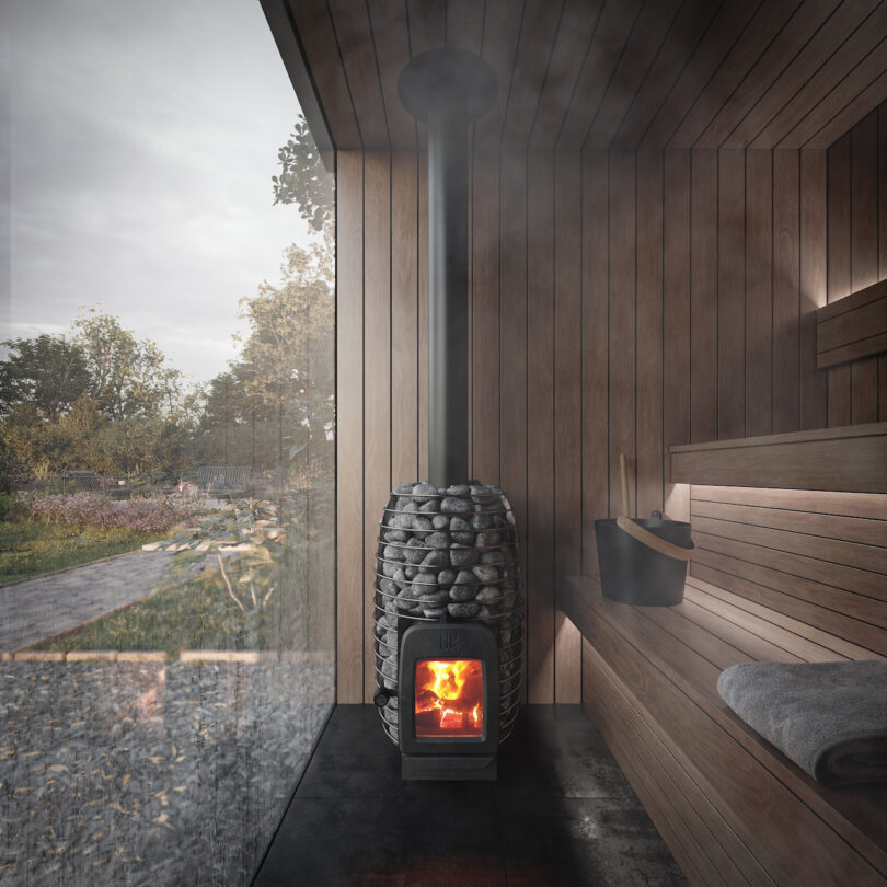 A wooden sauna interior featuring a stone-covered stove with a burning fire. A towel and bucket sit on the bench. A large glass window provides a view of the outdoor landscape