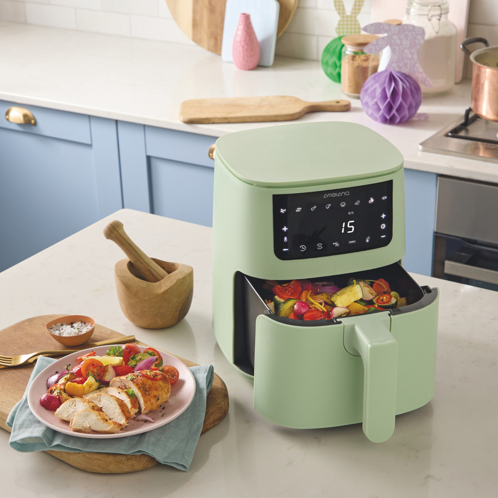 Aldi green air fryer on a kitchen island. It's drawer is filled with cooked veg and next to it, is a plate of roast chicken and vegetables.