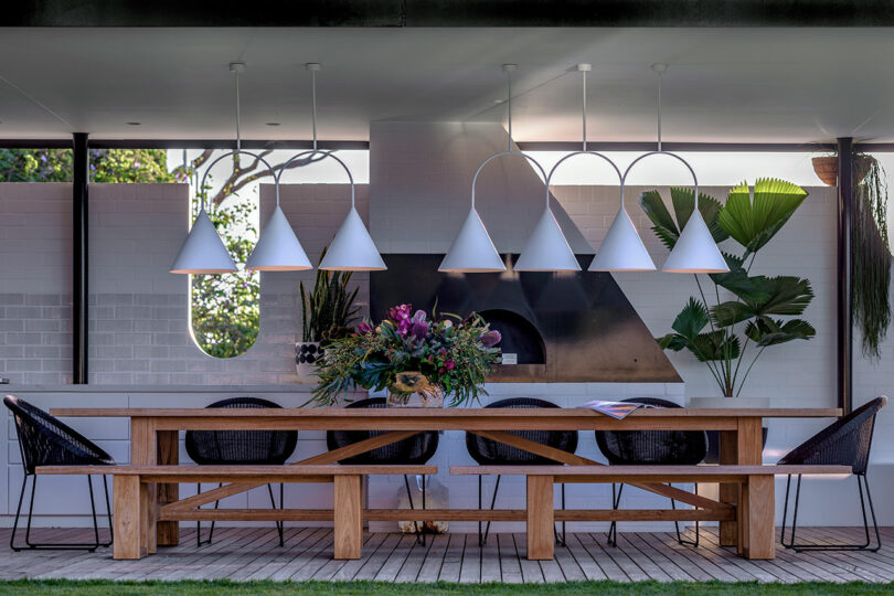 A modern, LA Cool outdoor dining area features a wooden table, black chairs, and white pendant lights. A stylish flower arrangement adorns the table, while large green plants create a chic backdrop.
