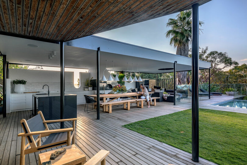 LA cool meets relaxation in this open-air modern patio. Featuring wooden decking, a chic dining area, stylish lounge chairs, and a glistening swimming pool surrounded by lush trees. A laid-back vibe is evident as a person sits chilled at the dining table.