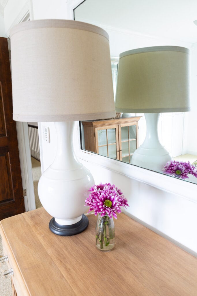 lamp and flowers on a wood cabinet in a bedroom with another wood pieces reflected in mirror.