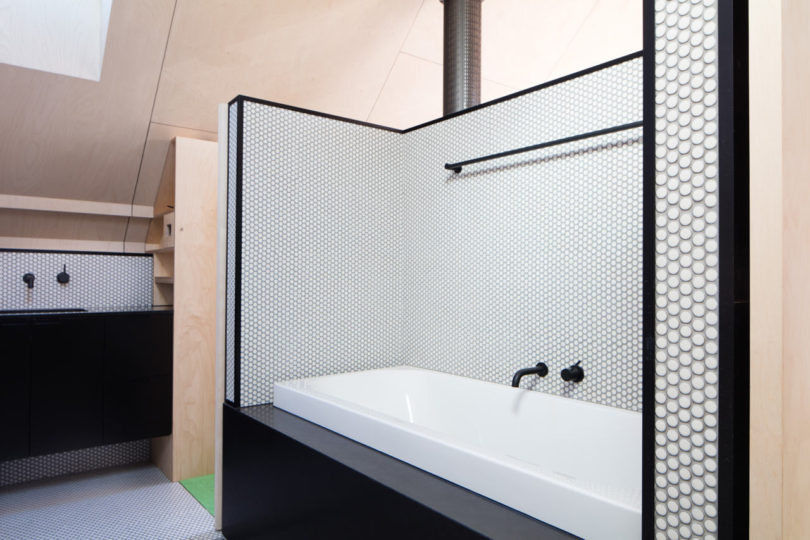 Modern bathroom in a triangular-shaped house with a white bathtub, black fixtures, and walls covered in small hexagonal white tiles bordered with black. Sink and cabinets are visible in the background.