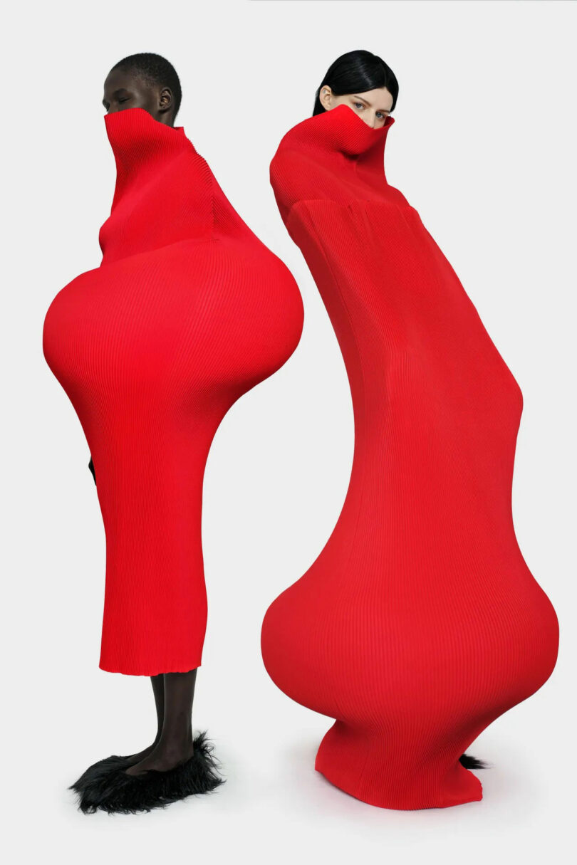 Two models wear exaggerated, form-fitting red dresses with extreme hourglass shapes. One dress covers the lower face; both outfits distort the natural body silhouette against a plain background.