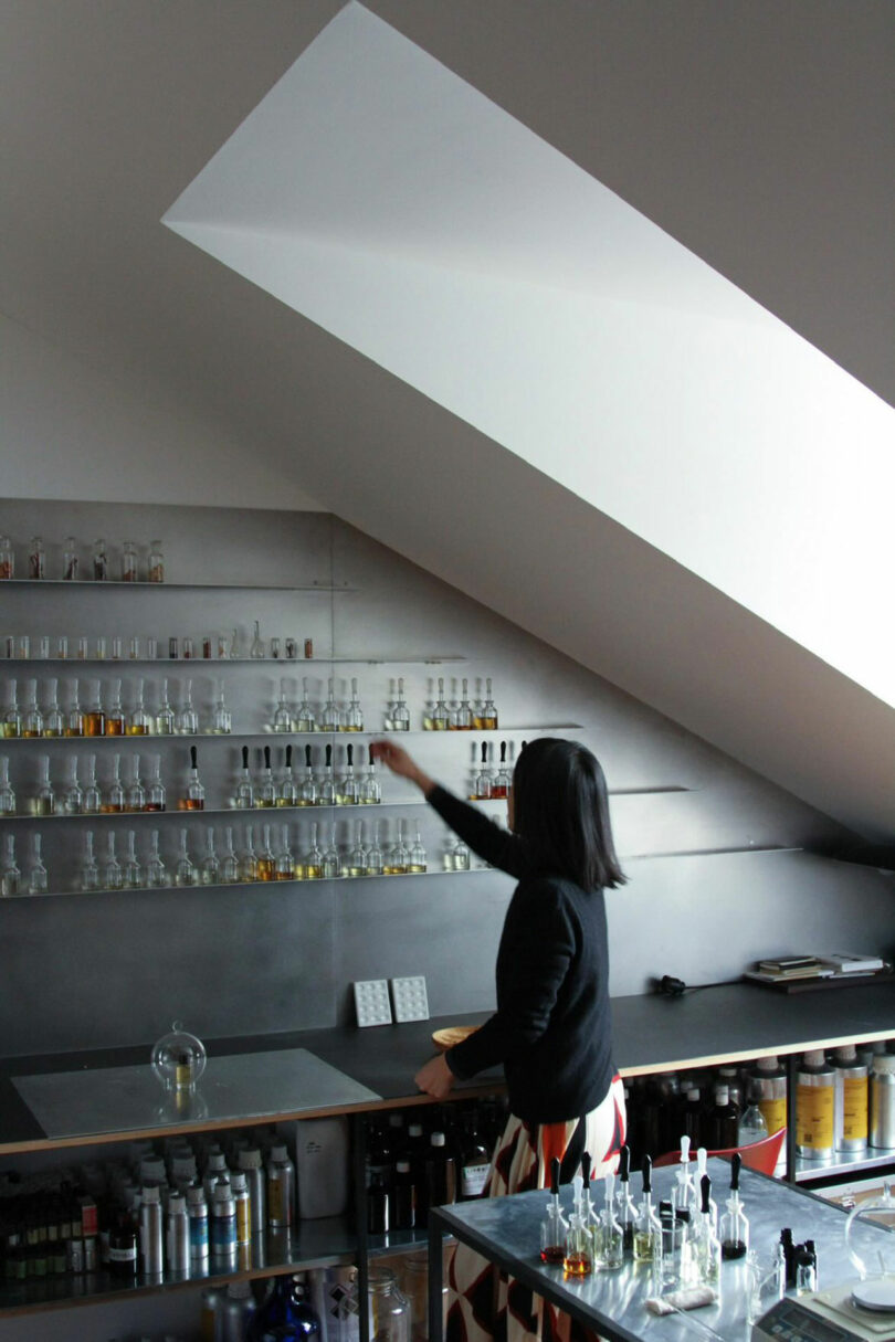 A person stands in a modern lab or studio, reaching for a glass bottle on a shelf filled with various bottles, under a slanted ceiling.