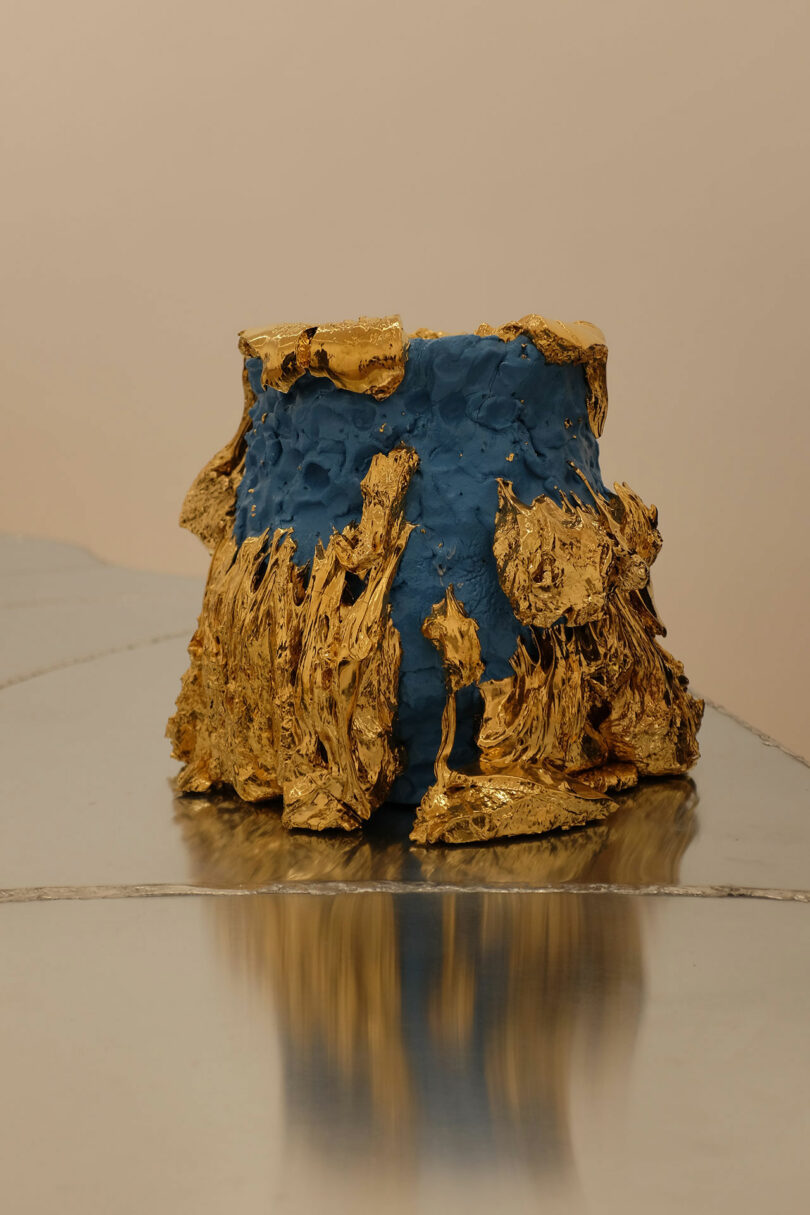 A blue ceramic vessel with textured, irregular gold accents on its top and base, displayed on a reflective metallic surface against a neutral background.