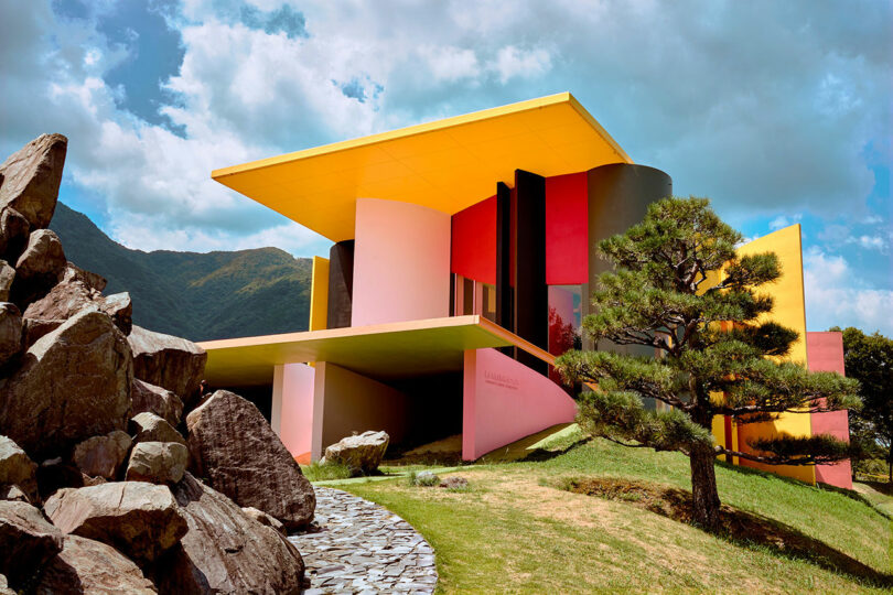 Modern house with bold geometric shapes and bright colors, including yellow, pink, black, and red, set in a landscaped yard with rocks and a tree under a partly cloudy sky.