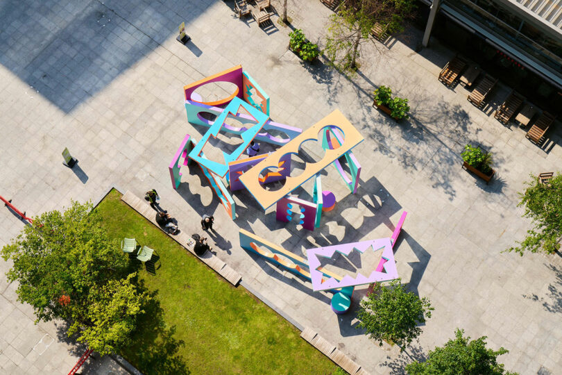Aerial view of a colorful geometric outdoor art installation on a paved plaza, surrounded by trees, grass, and people nearby.