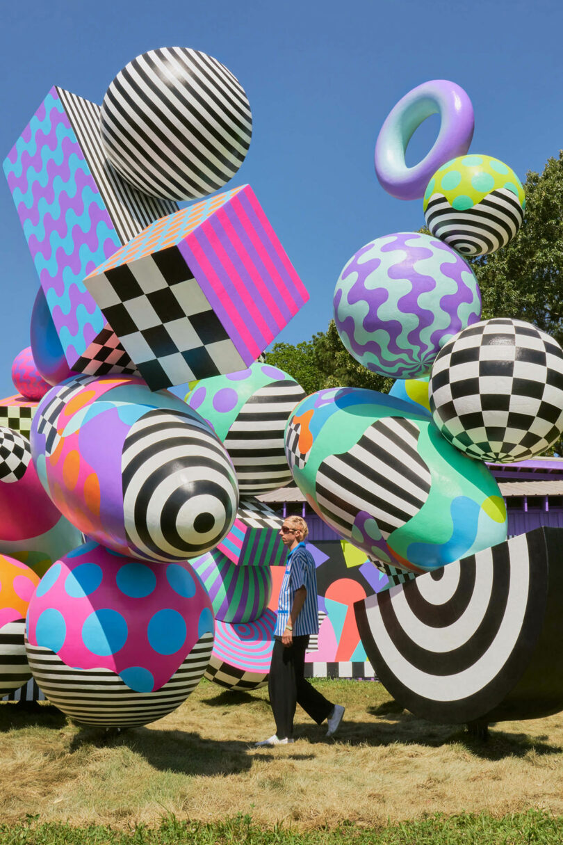 A person stands among large, colorful, geometric sculptures featuring stripes, polka dots, and checkerboard patterns on a sunny day.