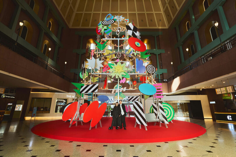 A large, colorful abstract sculpture with geometric shapes stands on a red carpet in the center of a spacious mall atrium, with two people posing in front.