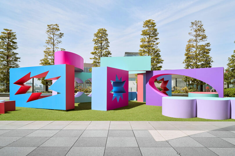 Colorful geometric sculptures with star-shaped cutouts are displayed on a grassy area, with trees and a building in the background under a clear sky.