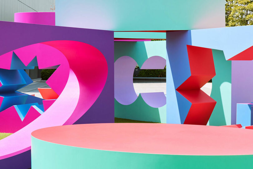 Colorful geometric sculptures with circular, rectangular, and jagged cutouts arranged outdoors, featuring vibrant pink, purple, blue, green, and red shapes.