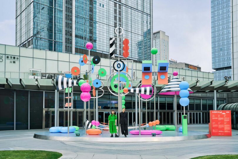 Two people stand in front of a colorful, abstract outdoor art installation with geometric shapes and large city buildings in the background.