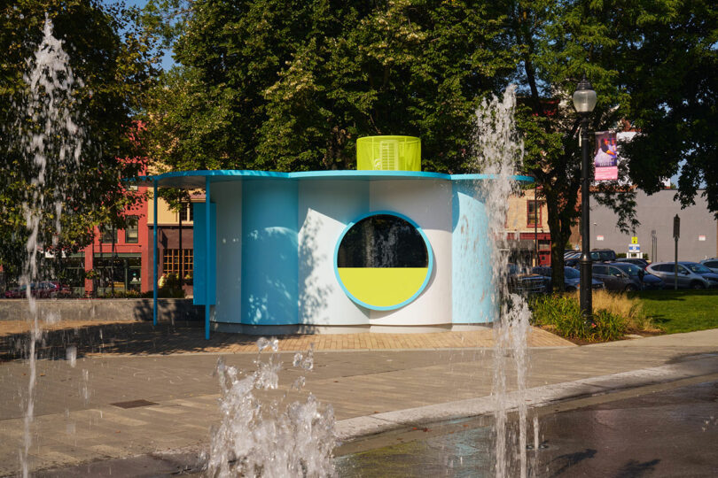 Small, round building with blue and yellow accents stands near a paved walkway, surrounded by trees and water fountains in a city park.