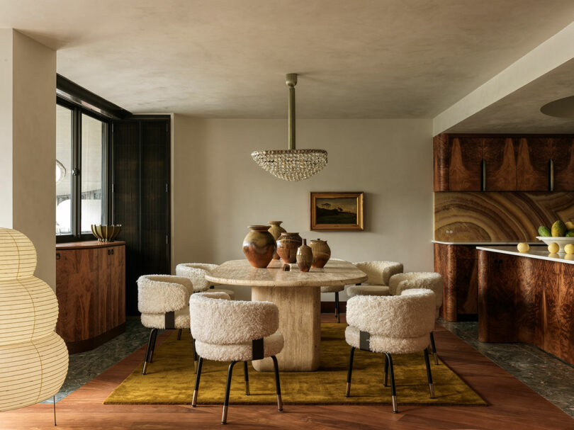 A modern dining room with a round stone table, six white textured chairs, ceramic vases, a crystal chandelier, wood cabinetry, and a yellow rug on a herringbone floor.