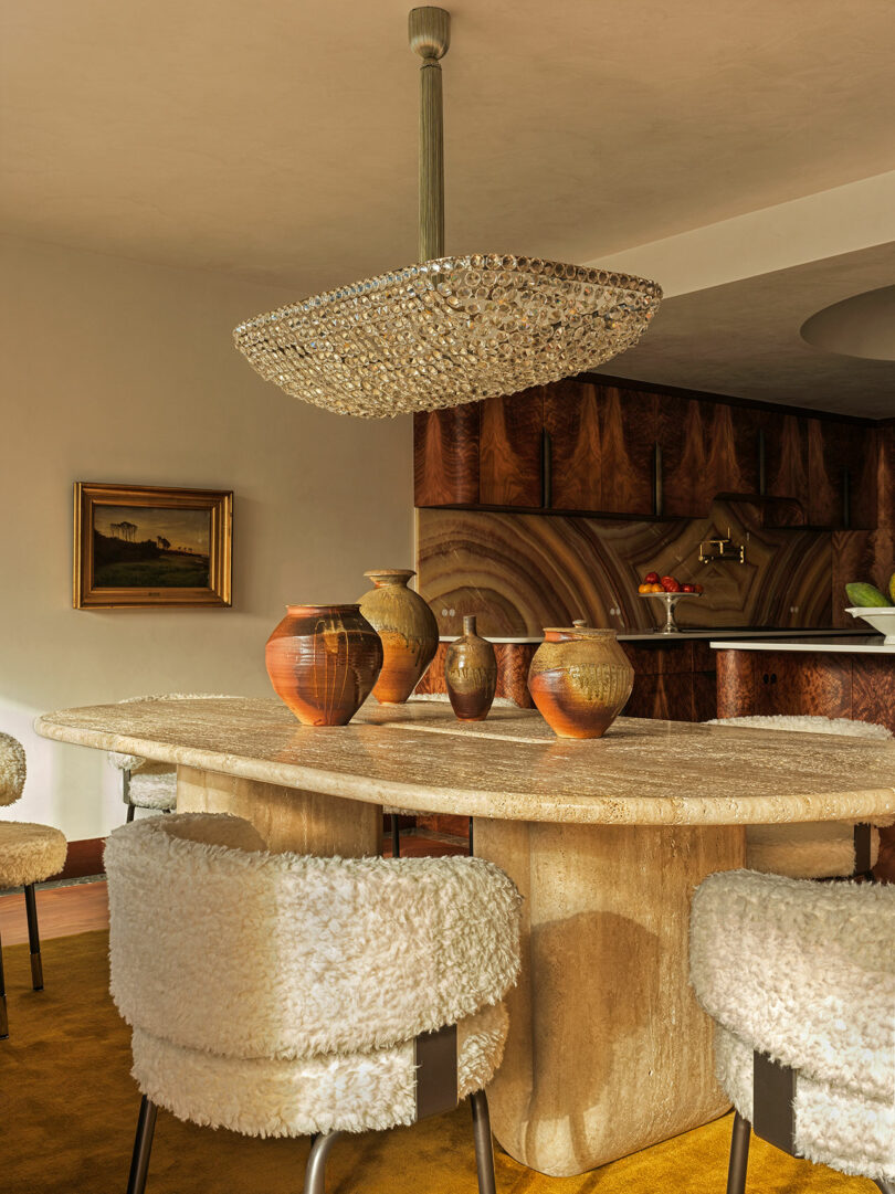 A dining area with a textured stone table, ceramic vases, boucle-covered chairs, a large crystal chandelier, and a wood-paneled kitchen in the background.