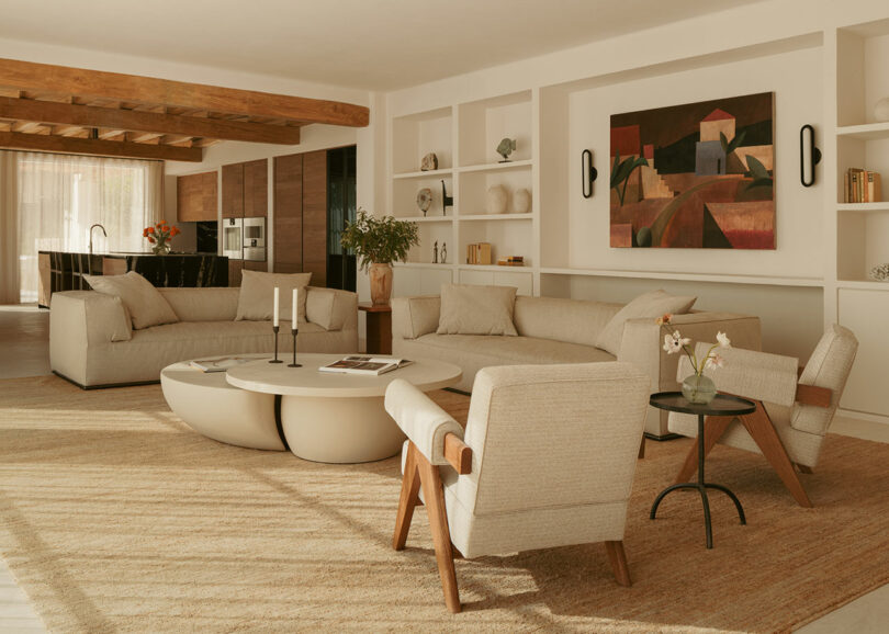 Modern living room with beige sofas, a round coffee table, wooden armchairs, built-in shelves, and a geometric art piece on the wall, featuring natural light and neutral tones.