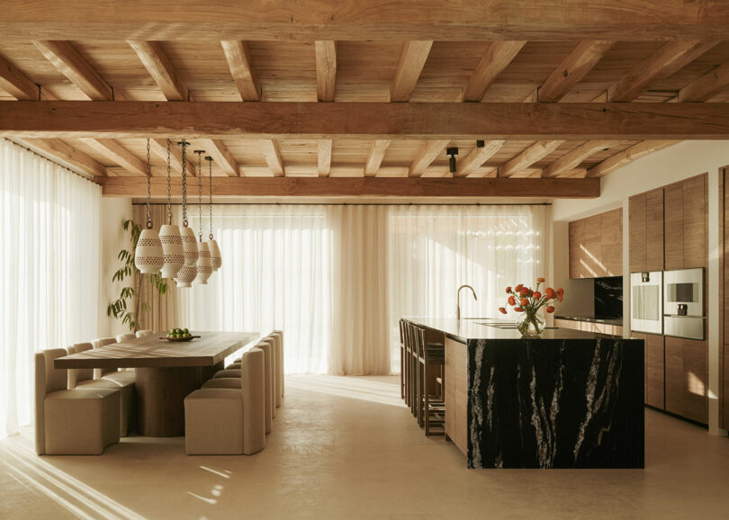 Modern kitchen and dining area with wooden ceiling, marble island, built-in appliances, pendant lights, and large windows with sheer curtains allowing natural light.