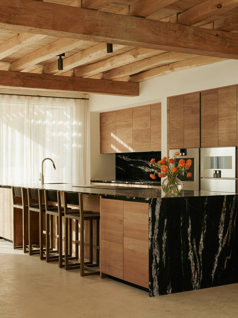 Modern kitchen with wooden cabinets, black marble countertops, a kitchen island with bar stools, flowers in a vase, and exposed wooden ceiling beams. Natural light filters through sheer curtains.
