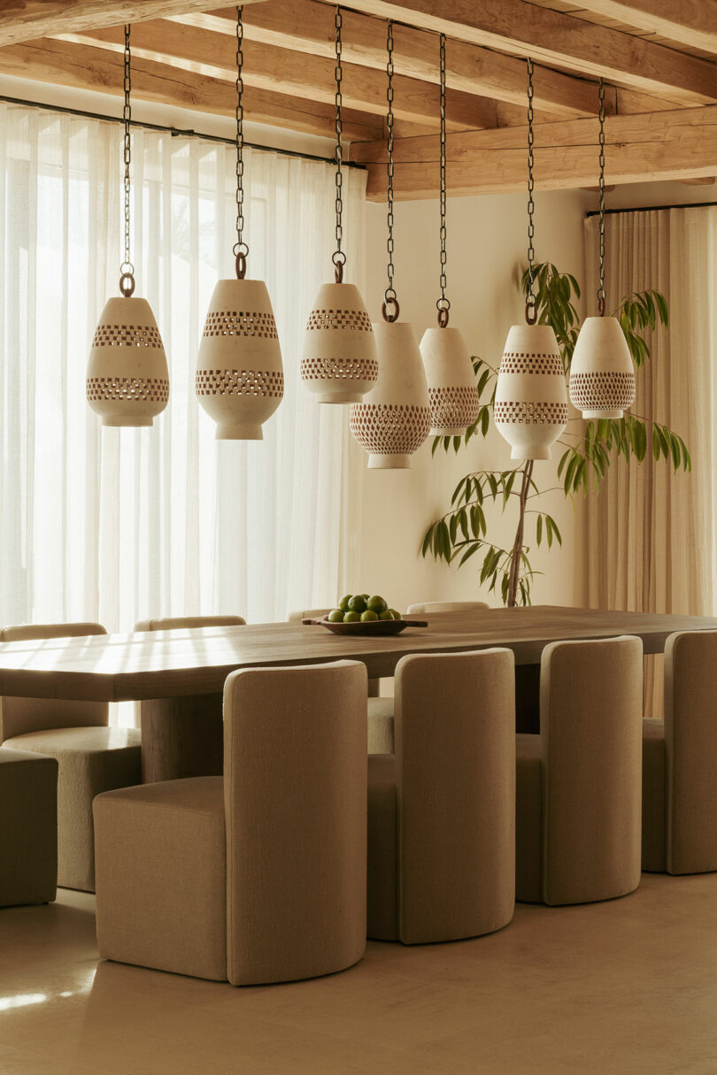 Modern dining room with a long wooden table, beige upholstered chairs, ceramic pendant lights, sheer curtains, and a potted plant in the corner.