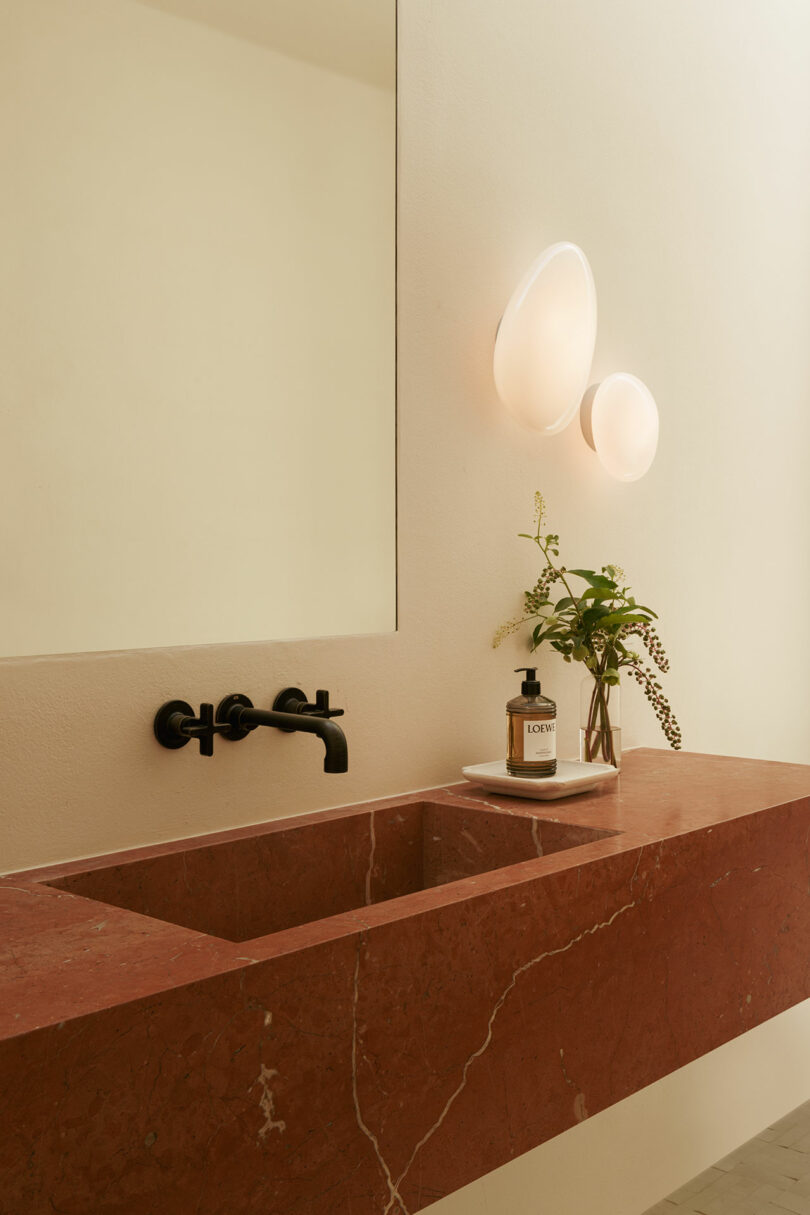 Minimalist bathroom with a red marble sink, black wall-mounted faucet, large mirror, modern wall lights, and a small vase with greenery beside a soap dispenser.