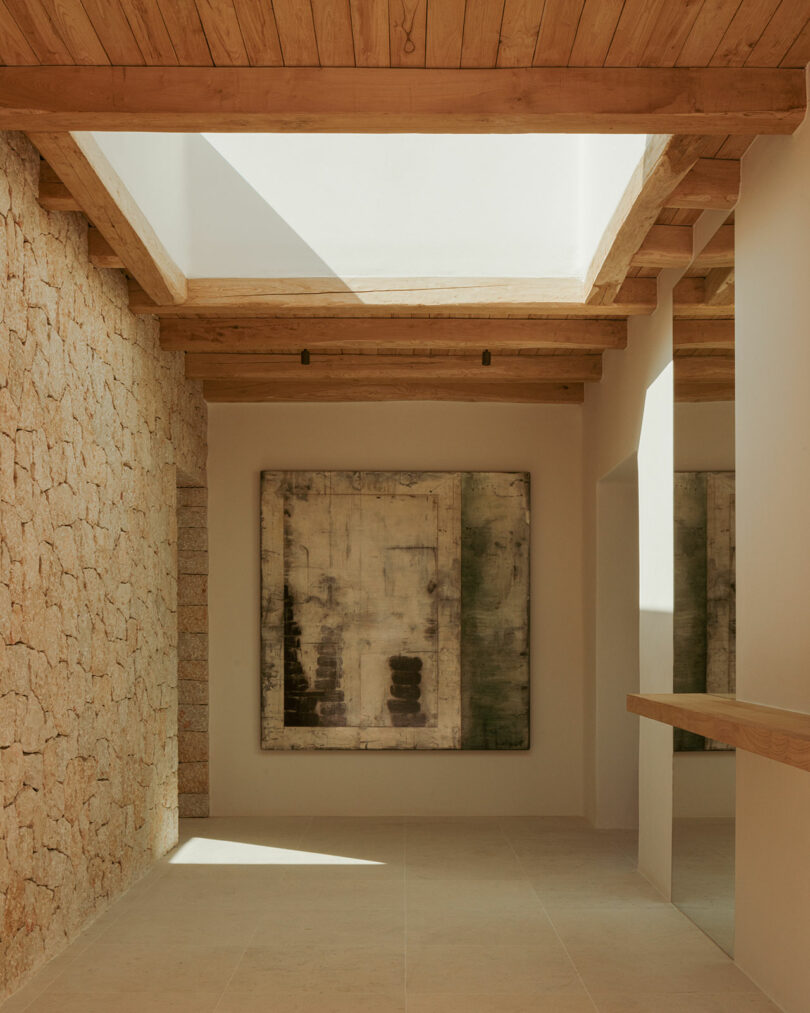 A hallway with stone and plaster walls, wooden ceiling beams, skylight, large abstract painting, and minimal natural light.