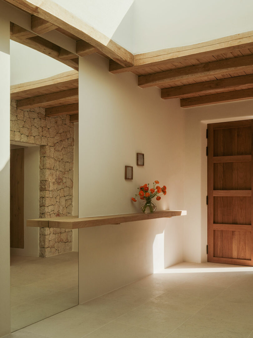 Minimalist entryway with a wooden door, stone wall, beige tiles, floating wooden shelf, and a glass vase of orange flowers. Two small square frames hang above the shelf.