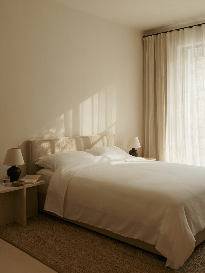 A minimalist bedroom with a neatly made bed, two bedside tables with matching lamps, beige walls, sheer curtains, and soft natural light streaming in.